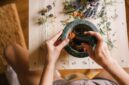 Seorang perempuan Melayu sedang meracik ramuan herbal tradisional, menunjukkan dedikasi dalam melestarikan kearifan lokal untuk kesehatan dan kecantikan alami. Rempah-rempah dan daun-daunan segar tampak tersusun rapi, siap diolah menjadi eliksir kehidupan.