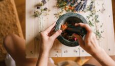 Seorang perempuan Melayu sedang meracik ramuan herbal tradisional, menunjukkan dedikasi dalam melestarikan kearifan lokal untuk kesehatan dan kecantikan alami. Rempah-rempah dan daun-daunan segar tampak tersusun rapi, siap diolah menjadi eliksir kehidupan.