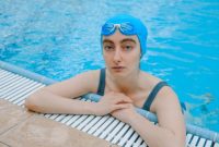Seorang wanita melakukan gerakan aqua aerobic dengan riang di dalam kolam renang, menunjukkan bagaimana olahraga air bisa menjadi aktivitas kebugaran yang menyenangkan dan menyegarkan. Latihan di dalam air mengurangi beban pada sendi sekaligus memberikan resistensi alami untuk melatih otot secara efektif.
