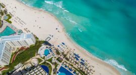 Pemandangan udara menakjubkan dari pantai pasir putih yang membentang luas, dihiasi perpaduan biru laut jernih dan hijaunya vegetasi tropis di pesisir. Sebuah surga tersembunyi yang menawarkan kedamaian dan keindahan alami.